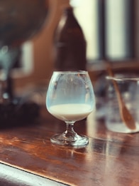 A half-filled snifter glass is placed on a wooden table, containing a light, milky liquid. The background is softly blurred, revealing a large bottle and some utensils, with light streaming through a window.