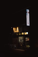 A dimly lit entrance to a guest house is visible, with a sign that reads 'Khaosan Kyoto'. Warm lights illuminate the interior, revealing a cozy atmosphere. Potted plants are placed in front of the entrance, adding a touch of greenery.