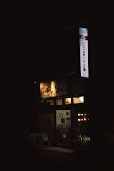 A dimly lit entrance to a guest house is visible, with a sign that reads 'Khaosan Kyoto'. Warm lights illuminate the interior, revealing a cozy atmosphere. Potted plants are placed in front of the entrance, adding a touch of greenery.