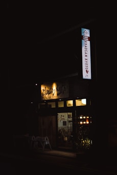A dimly lit entrance to a guest house is visible, with a sign that reads 'Khaosan Kyoto'. Warm lights illuminate the interior, revealing a cozy atmosphere. Potted plants are placed in front of the entrance, adding a touch of greenery.