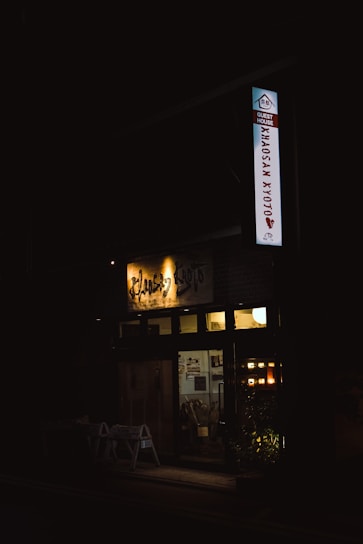 A dimly lit entrance to a guest house is visible, with a sign that reads 'Khaosan Kyoto'. Warm lights illuminate the interior, revealing a cozy atmosphere. Potted plants are placed in front of the entrance, adding a touch of greenery.