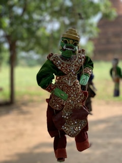 A traditional puppet dressed in ornate clothing featuring intricate patterns and embellishments, with a green face and detailed headdress, suspended in mid-air against a blurred natural background.