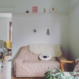 A cozy living room corner with a light brown leather couch adorned with a fluffy white throw and an eye-shaped pillow. A plant sits in the foreground on a small table. The walls feature minimalistic decor including a magazine, a small photo, and a wall hanging. An adjacent room with a yellow chair can be seen through a doorway.