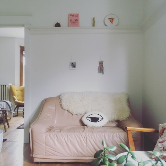 A cozy living room corner with a light brown leather couch adorned with a fluffy white throw and an eye-shaped pillow. A plant sits in the foreground on a small table. The walls feature minimalistic decor including a magazine, a small photo, and a wall hanging. An adjacent room with a yellow chair can be seen through a doorway.