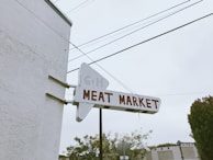 A vintage sign with the words 'Meat Market' in bold red letters against a white background is affixed to the side of a building. The sign is shaped like an arrow, pointing right, with some faint lettering visible above. The scene includes overhead power lines and cloud-covered sky in the background, surrounded by trees and another building.