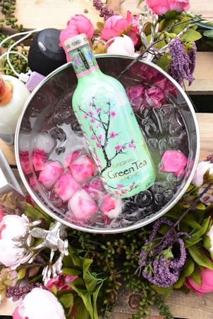 A vibrant arrangement showcasing a green tea bottle with cherry blossom artwork, submerged in ice with pink rose petals. The scene is framed by various pink and purple flowers and greenery, exuding a fresh and elegant feel.