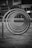 Several circular metal loops are aligned in a row, creating a tunnel effect in a modern urban setting. The background includes buildings with signage and neatly trimmed shrubs.