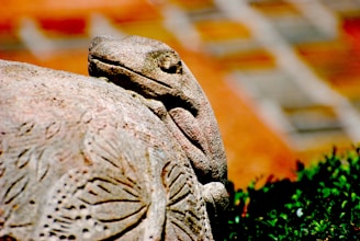 A stone sculpture of a lizard resting on a rock, intricately carved with patterns resembling nature. The background includes a blurred mix of earthy tones.