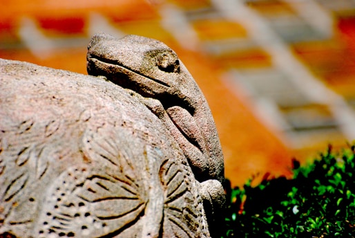 A stone sculpture of a lizard resting on a rock, intricately carved with patterns resembling nature. The background includes a blurred mix of earthy tones.