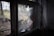 A window with shattered glass overlooking a blurred outdoor scene with trees and soft natural light. The broken glass panes are jagged with some pieces missing.