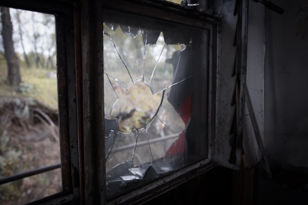 A window with shattered glass overlooking a blurred outdoor scene with trees and soft natural light. The broken glass panes are jagged with some pieces missing.