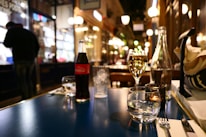A cozy, warmly lit restaurant scene with a focus on a table set with a bottle of Coca-Cola, wine, water glasses, cutlery, and a napkin. The background reveals a blurred interior with soft glowing lights and a person standing near a counter.