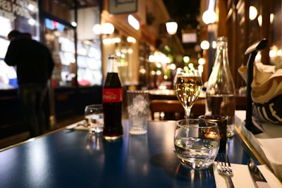 A cozy, warmly lit restaurant scene with a focus on a table set with a bottle of Coca-Cola, wine, water glasses, cutlery, and a napkin. The background reveals a blurred interior with soft glowing lights and a person standing near a counter.