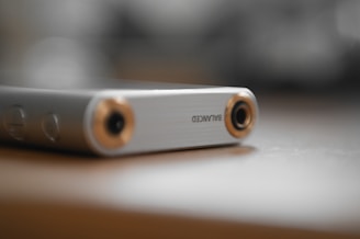 A close-up of a sleek, metallic audio device with a balanced input/output jack, featuring a brushed silver finish with gold accents. It is resting on a smooth, light-colored surface with a blurred background, creating a soft and elegant appearance.