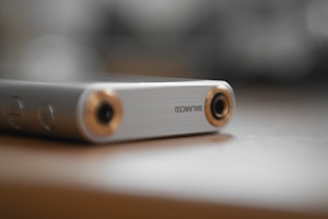 A close-up of a sleek, metallic audio device with a balanced input/output jack, featuring a brushed silver finish with gold accents. It is resting on a smooth, light-colored surface with a blurred background, creating a soft and elegant appearance.