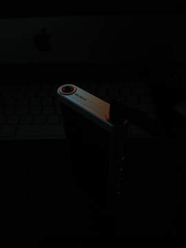 A darkly lit image featuring a close-up view of an electronic device. The device appears to be a rectangular audio interface or amplifier with a visible headphone jack. In the background, a partially visible keyboard and a faint Apple logo are discernible, suggesting a computer workspace setting.