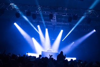 A DJ stands at a booth under a spotlight on a stage. Bright blue and white lights illuminate the scene, casting beams across the room. A crowd of people is visible in the foreground, silhouetted against the stage lighting.