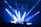 A DJ stands at a booth under a spotlight on a stage. Bright blue and white lights illuminate the scene, casting beams across the room. A crowd of people is visible in the foreground, silhouetted against the stage lighting.