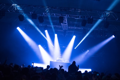 A DJ stands at a booth under a spotlight on a stage. Bright blue and white lights illuminate the scene, casting beams across the room. A crowd of people is visible in the foreground, silhouetted against the stage lighting.