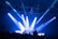 A DJ stands at a booth under a spotlight on a stage. Bright blue and white lights illuminate the scene, casting beams across the room. A crowd of people is visible in the foreground, silhouetted against the stage lighting.