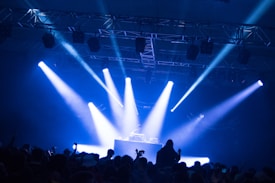 A DJ stands at a booth under a spotlight on a stage. Bright blue and white lights illuminate the scene, casting beams across the room. A crowd of people is visible in the foreground, silhouetted against the stage lighting.