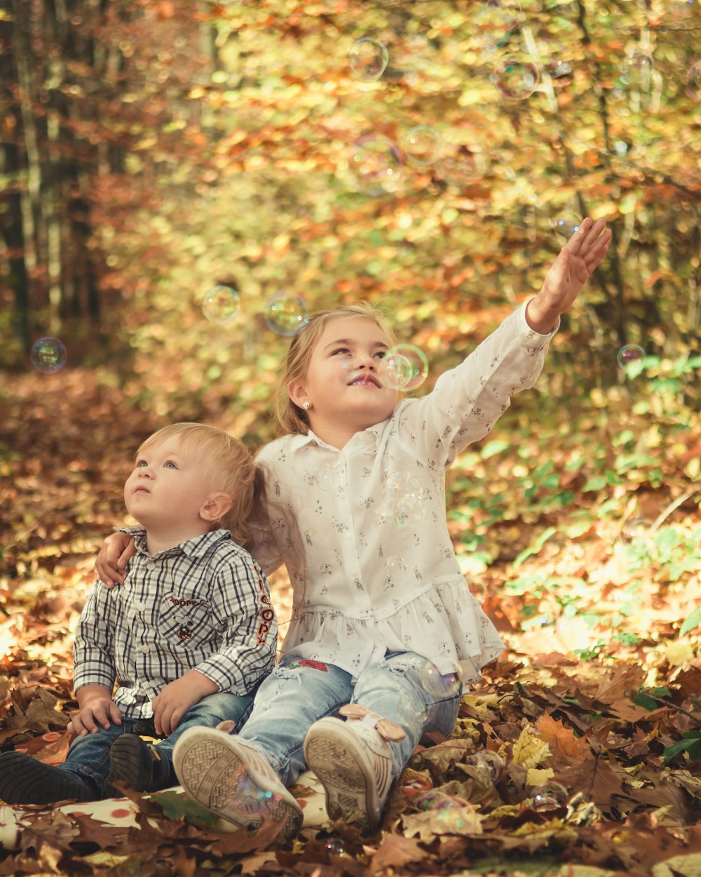 smiling girl sitting on ground near child looking up while reaching bubbles surrounded with tall and orange trees during daytime