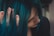 A person with dark hair dyed in vibrant teal is partially covering their face with their hand. The fingers have a natural manicure and the person wears dark lipstick, creating a contrasting look. The hair has a silky texture and catches the light subtly.