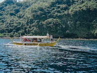 A small boat with people wearing life jackets is moving across a body of water. The boat, with its distinct outriggers, is typical of traditional watercraft found in Southeast Asian regions. In the background, a lush, densely forested island provides a vibrant green contrast against the deep blue of the water.