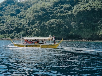 A small boat with people wearing life jackets is moving across a body of water. The boat, with its distinct outriggers, is typical of traditional watercraft found in Southeast Asian regions. In the background, a lush, densely forested island provides a vibrant green contrast against the deep blue of the water.