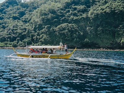 A small boat with people wearing life jackets is moving across a body of water. The boat, with its distinct outriggers, is typical of traditional watercraft found in Southeast Asian regions. In the background, a lush, densely forested island provides a vibrant green contrast against the deep blue of the water.