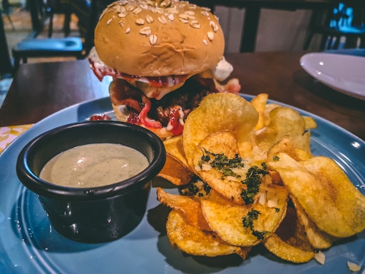 A plate contains a burger with a bun topped with oats, along with crispy chips garnished with herbs. A small black bowl of dipping sauce is placed next to the chips, all on a blue plate set on a wooden table.