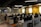 A modern office space with rows of empty desks equipped with office chairs and computer monitors. The walls are painted yellow and gray, with crossword-like text featuring brand names. The ceiling lights are linear and futuristic. Potted plants adorn the desks, adding a touch of greenery.