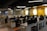 A modern office space with rows of empty desks equipped with office chairs and computer monitors. The walls are painted yellow and gray, with crossword-like text featuring brand names. The ceiling lights are linear and futuristic. Potted plants adorn the desks, adding a touch of greenery.