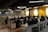 A modern office space with rows of empty desks equipped with office chairs and computer monitors. The walls are painted yellow and gray, with crossword-like text featuring brand names. The ceiling lights are linear and futuristic. Potted plants adorn the desks, adding a touch of greenery.