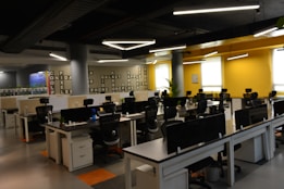 A modern office space with rows of empty desks equipped with office chairs and computer monitors. The walls are painted yellow and gray, with crossword-like text featuring brand names. The ceiling lights are linear and futuristic. Potted plants adorn the desks, adding a touch of greenery.