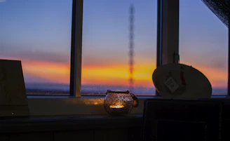 A cozy candlelit corner with a softly glowing Cyprus Candles jar beside a window showing a sunlit Mediterranean landscape.