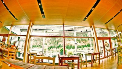 A cozy restaurant interior featuring large windows with a view of a lush, wooded landscape. The room has a warm wooden ceiling with modern lighting fixtures. Various tables and chairs are neatly arranged, some set for dining.