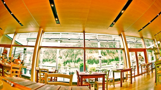 A cozy restaurant interior featuring large windows with a view of a lush, wooded landscape. The room has a warm wooden ceiling with modern lighting fixtures. Various tables and chairs are neatly arranged, some set for dining.