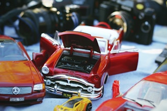 A collection of model cars, including a red classic car with open doors and hood, revealing the engine. There are also some cameras in the background, creating a sense of a vintage or hobbyist setting.
