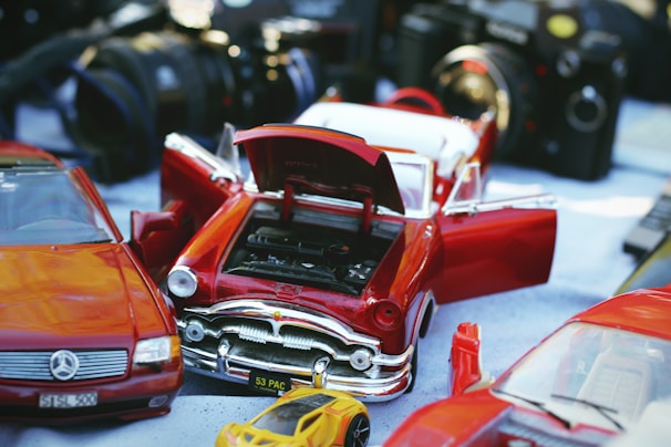 A collection of model cars, including a red classic car with open doors and hood, revealing the engine. There are also some cameras in the background, creating a sense of a vintage or hobbyist setting.