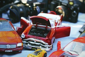 A collection of model cars, including a red classic car with open doors and hood, revealing the engine. There are also some cameras in the background, creating a sense of a vintage or hobbyist setting.