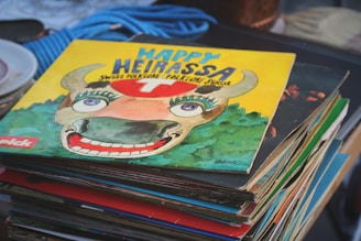 A stack of vinyl records is topped by an album with bright, vivid artwork. The top record's cover features a whimsical illustration of a bull with large eyes and a broad smile, set against a yellow background. The text on the cover reads 'Happy Heirassa' and includes the phrase 'Swiss Folklore / Folklore Suisse'. Surrounding the image are other record covers, slightly visible beneath it.
