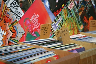 An assortment of vinyl records displayed in cardboard boxes at a market stall. The records are organized into categories with handwritten labels indicating various music genres such as soul, jazz, rock, and pop. Visible album covers feature vibrant artwork and typography, including artists like King Curtis.