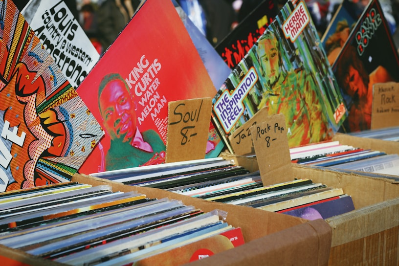 An assortment of vinyl records displayed in cardboard boxes at a market stall. The records are organized into categories with handwritten labels indicating various music genres such as soul, jazz, rock, and pop. Visible album covers feature vibrant artwork and typography, including artists like King Curtis.