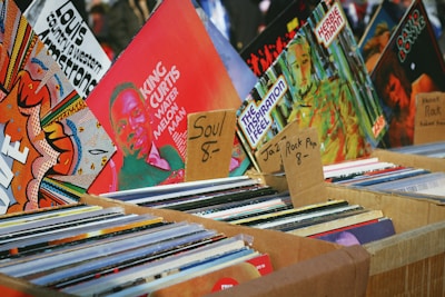 An assortment of vinyl records displayed in cardboard boxes at a market stall. The records are organized into categories with handwritten labels indicating various music genres such as soul, jazz, rock, and pop. Visible album covers feature vibrant artwork and typography, including artists like King Curtis.