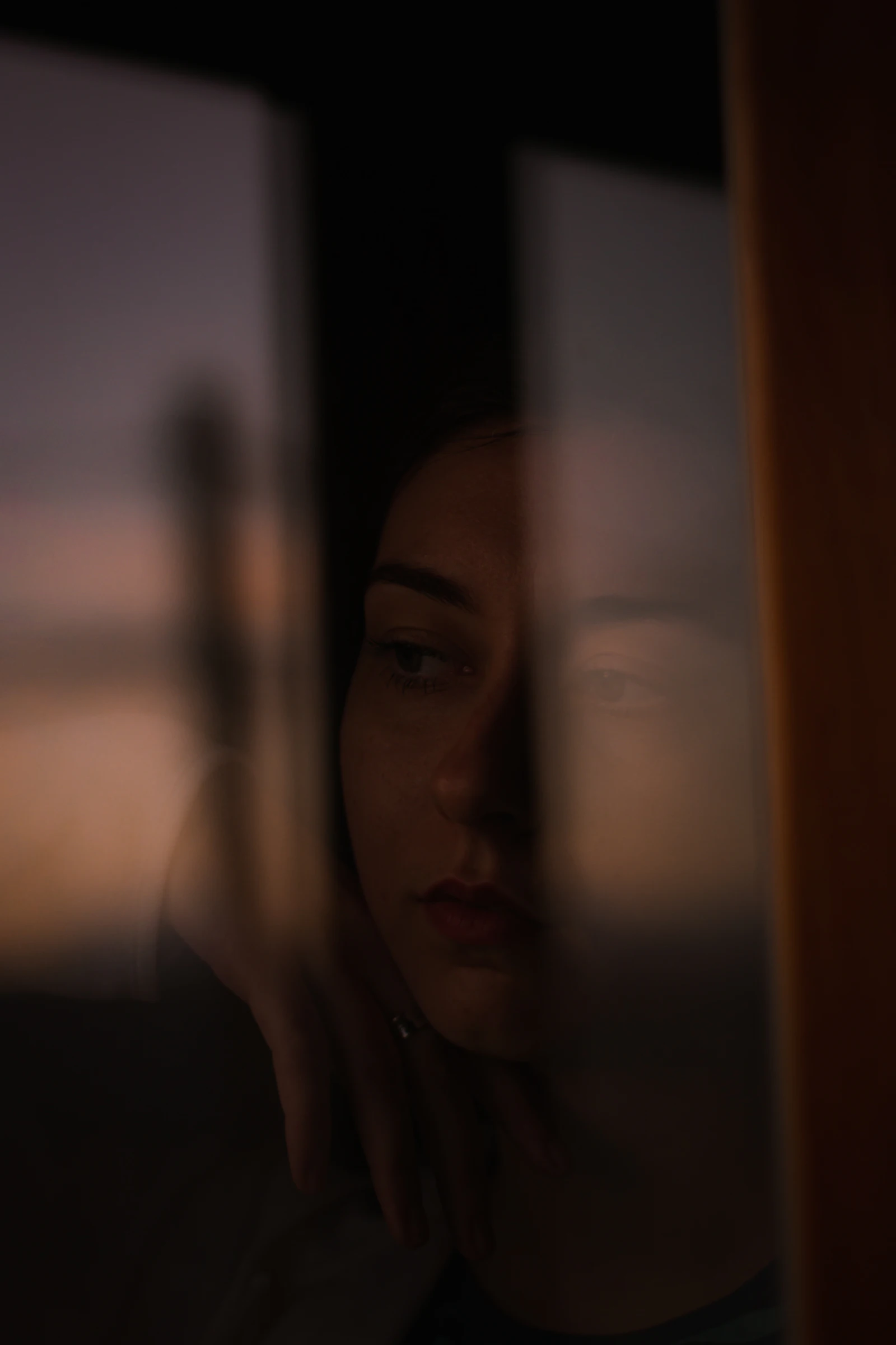 A young woman pictured at twilight through a reflected window, sunset glow on her face, soft flash, the kind of photograph that ambient-folk records ask for.