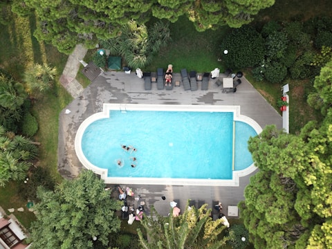 An aerial view of a swimming pool surrounded by wooden decking and lush green trees. Several people are enjoying the pool, while others relax on lounge chairs nearby. The scene is framed by dense foliage and vibrant greenery, adding to the secluded and tranquil atmosphere.