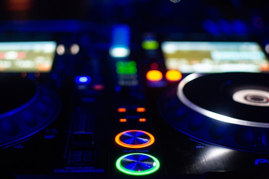 A close-up view of a DJ mixer with colorful LED lights illuminating the control buttons and components. The scene is set in a dimly lit environment, highlighting the vibrant colors on the mixer.