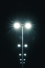 Several bright streetlights line up in the darkness, their light beams prominently illuminating the black night sky. The lights create a symmetrical pattern with glowing flares extending outward.