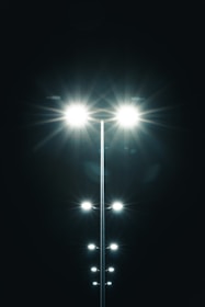 Several bright streetlights line up in the darkness, their light beams prominently illuminating the black night sky. The lights create a symmetrical pattern with glowing flares extending outward.
